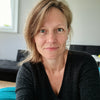 Frau in einem gut beleuchteten Raum, lächelt sanft in die Kamera, trägt einen schwarzen Pullover, im Hintergrund ein schwarzes Sofa und ein helles Fenster.