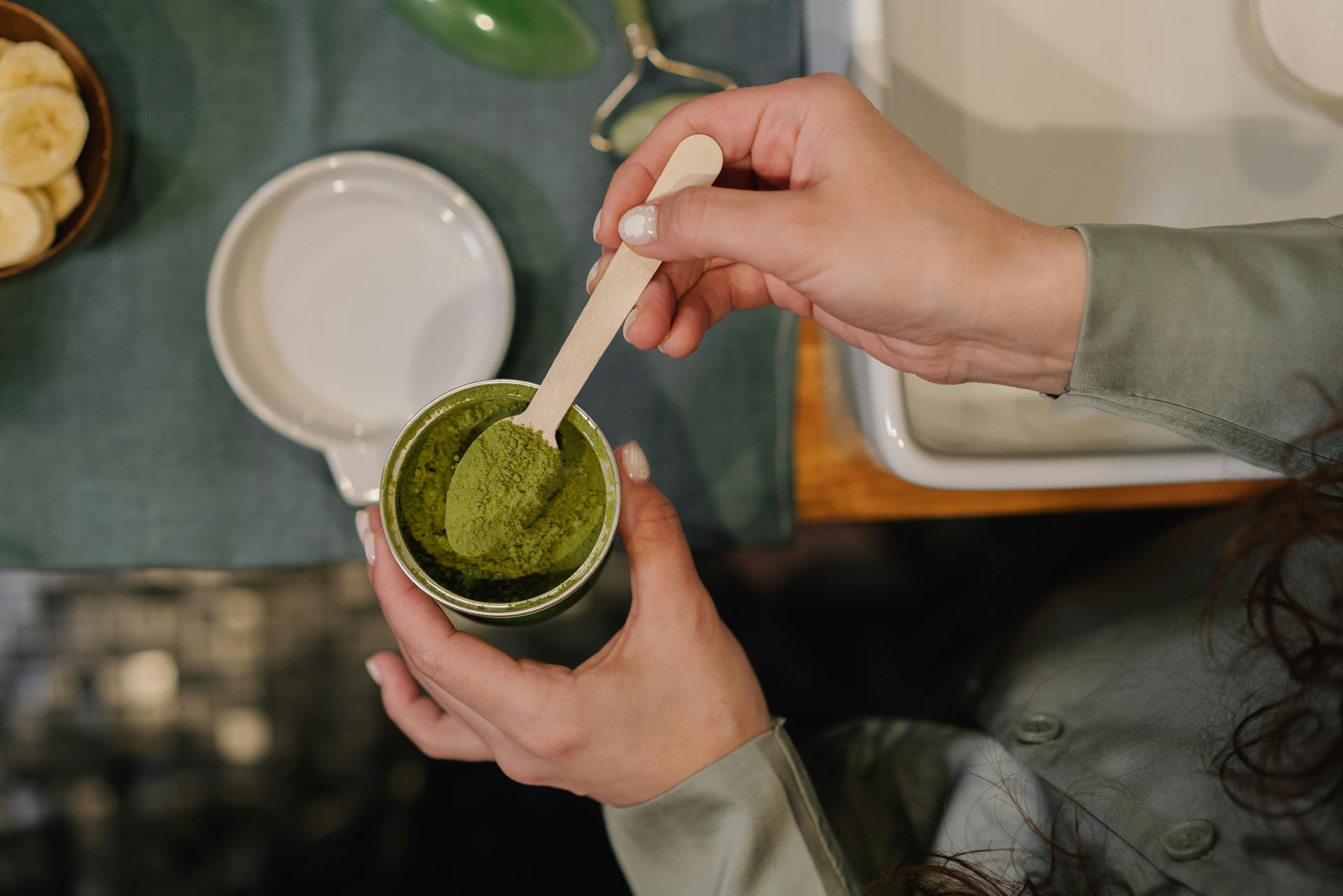 Frau holt mit einem Löffel Matcha Pulver aus Matcha-Dose. Im Hintergrund steht eine Matcha-Schale.